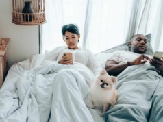 multiracial couple surfing mobile phones lying in bed with dog after awakening