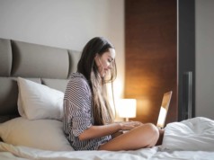 side view photo of smiling woman in a black and white striped top sitting on a bed while using a laptop