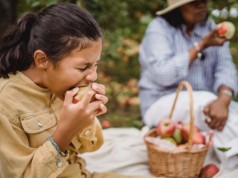 ethnic girl biting apple during picnic with mother