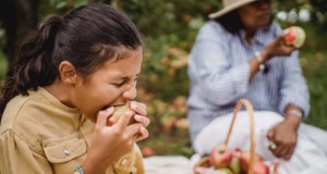 ethnic girl biting apple during picnic with mother