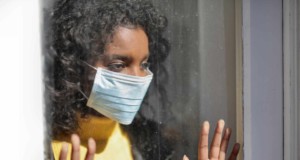 serious young ethnic lady in medical mask standing near window and looking away on street